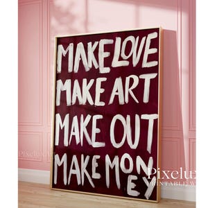 Puede incluir: Un cuadro rectangular en un marco dorado fino con un fondo color burdeos intenso y el texto pintado a mano en blanco: MAKE LOVE, MAKE ART, MAKE OUT, MAKE MONEY. Las letras tienen una textura visible de pinceladas.