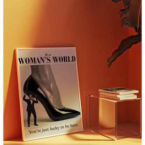 May include: A framed poster with the text "It's a WOMAN'S WORLD" and "You're just lucky to be here." The image features a high-heeled shoe and a man in a suit. A stack of books sits on a clear side table.