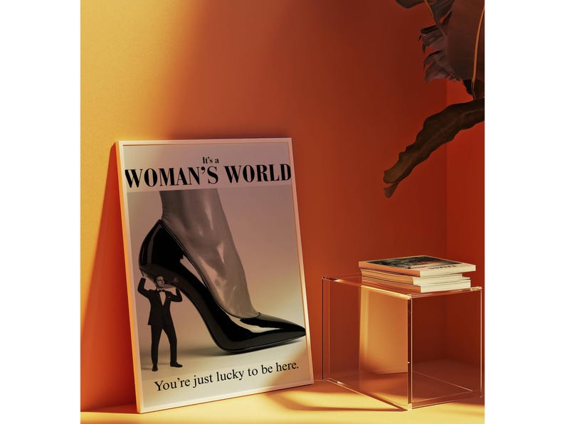 May include: A framed poster with the text "It's a WOMAN'S WORLD" and "You're just lucky to be here." The image features a high-heeled shoe and a man in a suit. A stack of books sits on a clear side table.