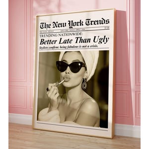 May include: A framed sepia-toned print featuring a woman applying lipstick, wearing sunglasses and a towel on her head. The print is designed to look like a newspaper, with the headline "The New York Trends" and the text "Better Late Than Ugly".