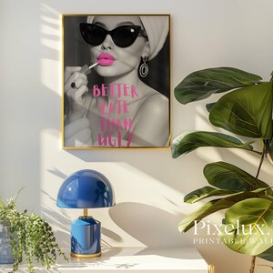 May include: A framed black and white print featuring a woman applying bright pink lipstick, wearing sunglasses and a headscarf. The text "BETTER LATE THAN UGLY" is in pink. A blue lamp and green plant are in the foreground.
