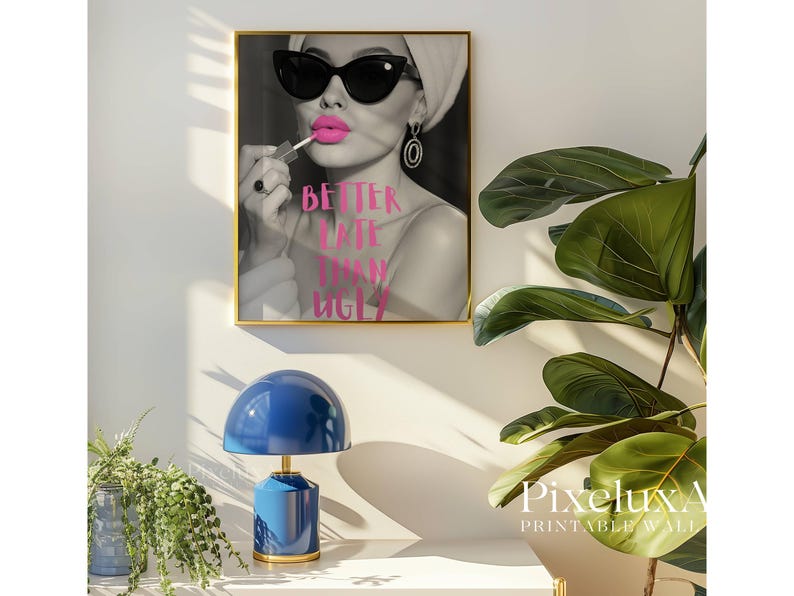 May include: A framed black and white print featuring a woman applying bright pink lipstick, wearing sunglasses and a headscarf. The text "BETTER LATE THAN UGLY" is in pink. A blue lamp and green plant are in the foreground.