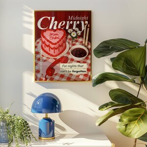 May include: A framed poster with the words "Midnight Cherry" and a heart-shaped cake illustration. Below, a blue table lamp with a rounded shade and a gold base sits on a white surface. A potted plant is to the right.