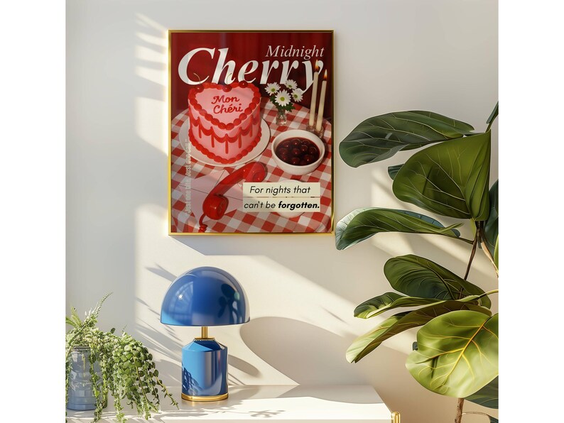 May include: A framed poster with the words "Midnight Cherry" and a heart-shaped cake illustration. Below, a blue table lamp with a rounded shade and a gold base sits on a white surface. A potted plant is to the right.