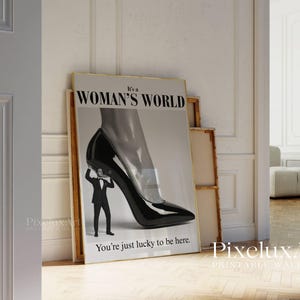 May include: A framed print with the text "It's a WOMAN'S WORLD" and "You're just lucky to be here." The artwork depicts a black high-heeled shoe with a man in a suit holding it up. The print is leaning against other framed pieces.