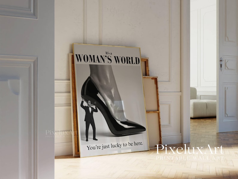 May include: A framed print with the text "It's a WOMAN'S WORLD" and "You're just lucky to be here." The artwork depicts a black high-heeled shoe with a man in a suit holding it up. The print is leaning against other framed pieces.