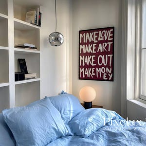 Puede incluir: Un cuadro rectangular enmarcado en negro con el texto blanco escrito a mano "MAKE LOVE MAKE ART MAKE OUT MAKE MONEY" sobre un fondo burdeos oscuro, expuesto sobre una cama con ropa de cama azul claro y una l&aacute;mpara de globo redonda.
