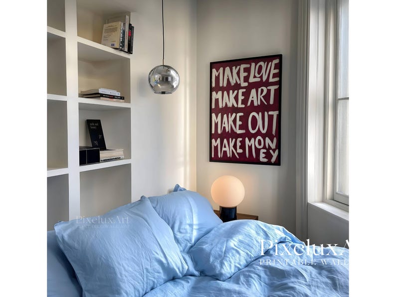 Puede incluir: Un cuadro rectangular enmarcado en negro con el texto blanco escrito a mano "MAKE LOVE MAKE ART MAKE OUT MAKE MONEY" sobre un fondo burdeos oscuro, expuesto sobre una cama con ropa de cama azul claro y una l&aacute;mpara de globo redonda.
