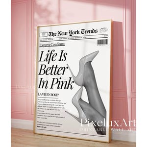 May include: A framed print featuring a newspaper-style design with the headline "Life Is Better In Pink." The image includes a grayscale photo of legs wearing high heels. The print is displayed against a pink wall.