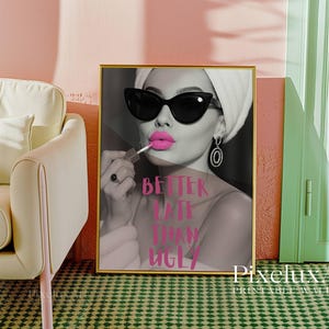May include: A framed print with a black and white image of a woman wearing sunglasses and applying pink lipstick. The text "BETTER LATE THAN UGLY" is in pink. The print is in a gold frame and is displayed in a room with a pink wall.