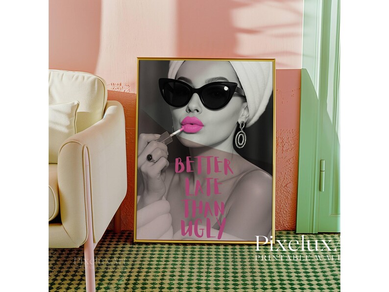 May include: A framed print with a black and white image of a woman wearing sunglasses and applying pink lipstick. The text "BETTER LATE THAN UGLY" is in pink. The print is in a gold frame and is displayed in a room with a pink wall.
