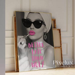 May include: Black and white print of a woman applying pink lipstick, wearing sunglasses and a headscarf. The text "BETTER LATE THAN UGLY" is in pink. The print is in a gold frame, leaning against a wall.