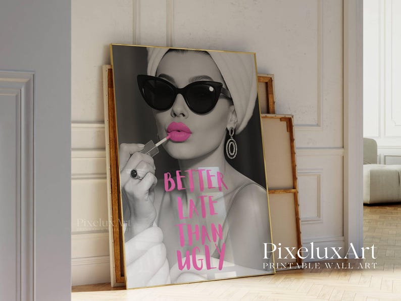 May include: Black and white print of a woman applying pink lipstick, wearing sunglasses and a headscarf. The text "BETTER LATE THAN UGLY" is in pink. The print is in a gold frame, leaning against a wall.