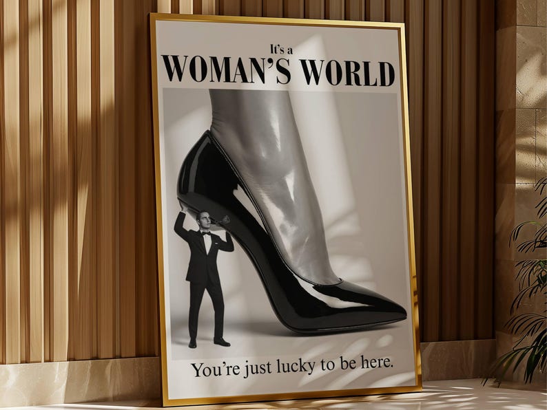 May include: Framed art print with a gold border. The image features a giant black high-heeled shoe with a man in a tuxedo holding it up. The text reads "It's a WOMAN'S WORLD" and "You're just lucky to be here."