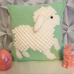 Könnte beinhalten: Ein grünes gehäkeltes Kissen mit einem weißen gehäkelten Lammdesign. Das Lamm hat ein schwarzes Auge und einen weißen, flauschigen Körper.