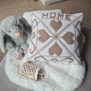 Könnte beinhalten: Ein weißes und braunes Häkelkissen mit dem Wort "HOME" in braunem Garn. Das Kissen liegt auf einem weißen Kunstfellteppich.