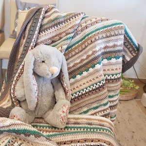 Op de afbeelding: Een gehaakt deken in tinten bruin, beige en groen met een witte rand. Een grijs pluche konijn zit op de deken.
