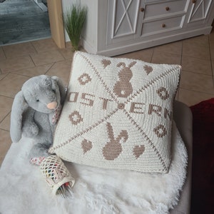 Könnte beinhalten: Ein gehäkeltes Kissen mit einem braunen Hasen und dem Wort "Ostern" in braunen Buchstaben. Das Kissen liegt auf einem weißen Kunstfellteppich.