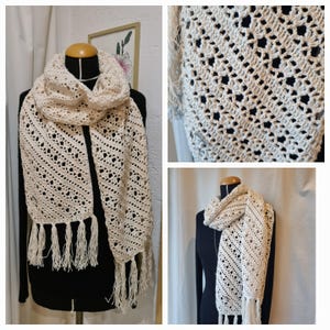 May include: A crocheted scarf with a delicate openwork pattern and fringe tassels. The scarf is cream coloured and is displayed on a mannequin.
