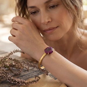 May include: A gold-toned cuff bracelet featuring a textured, hammered metal band and a single, large, irregular-shaped deep purple gemstone set in a prong setting. The bracelet is worn on the wrist, showcasing the contrast between the gold metal and stone.