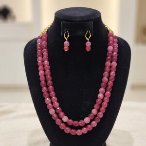 Pode incluir: Um colar de duas voltas com contas facetadas de cor rosa escuro com aspeto rachado e brincos pendentes a condizer. O conjunto de joalharia inclui peças e ganchos de metal dourado, com cada brinco composto por uma conta suspensa num fio.