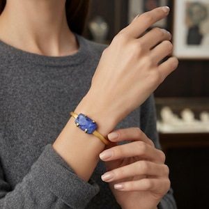 Puede incluir: Un brazalete de metal en tono dorado con una sola piedra preciosa azul rectangular y facetada, montada en cuatro garras. El brazalete tiene un acabado mate y liso, y se lleva alrededor de la muñeca, destacando la piedra de color azul intenso.