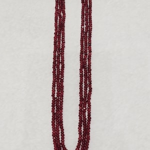 Può includere: Una collana a più fili composta da tre lunghe file di piccole perline sfaccettate color rosso scuro. I fili sono uniti nella parte superiore da due connettori in metallo dorato, creando un effetto a cascata a strati con perline uniformi.