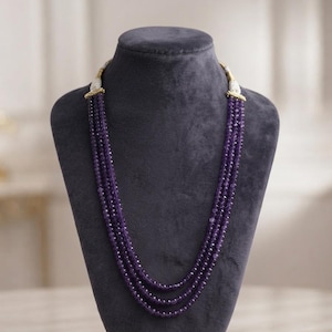 Puede incluir: Un collar de múltiples hilos con tres capas de cuentas de amatista de color púrpura intenso facetadas. Los hilos están conectados en la parte superior por detalles de metal dorado y cuentas blancas, creando un diseño en capas que cae en cascada.