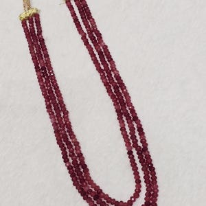 May include: A three-strand necklace featuring small, faceted, deep red beads. The strands are connected at the top by gold-toned metal caps with textured detailing, leading to a thin, light-coloured cord for fastening around the neck.