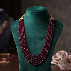 Puede incluir: Un collar de múltiples hilos con cinco filas de cuentas circulares facetadas de color rojo intenso. Los hilos están conectados en la parte superior por terminales metálicos dorados, ornamentados y texturizados, que aseguran las cuentas.