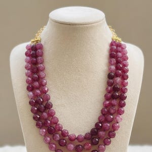 May include: A three-strand necklace featuring faceted, deep magenta and purple beads with a cracked texture. The strands are connected by gold-toned metal hardware at the top, creating a layered look with uniform, round, semi-translucent stone beads.