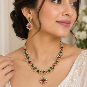 Peut inclure: Un ensemble de collier et boucles d'oreilles pendantes doré, orné de pierres en forme de goutte alternant entre le vert et le magenta. Les bijoux sont agrémentés de petites pierres blanches transparentes disposées en motif floral.