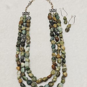 Puede incluir: Un collar de tres hilos con piedras pulidas de colores verdes y marrones, con espaciadores de metal plateado y cierre de cadena, acompañado de un par de pendientes a juego con ganchos plateados y detalles de piedra.