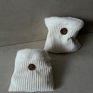 Könnte beinhalten: Zwei weiße Cord-Beutel mit flauschigem weißen Futter und einem Holzknopfverschluss.