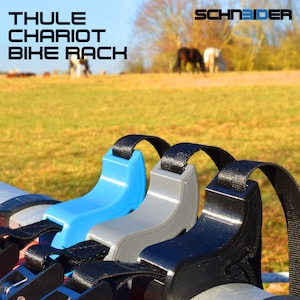 Peut inclure: Trois sangles de porte-vélos avec des supports en plastique de différentes couleurs. Les sangles sont noires et les supports sont bleus, gris et noirs. Les sangles sont fixées à une barre métallique argentée. Le texte "THULE CHARIOT BIKE RACK" est visible en arrière-plan.