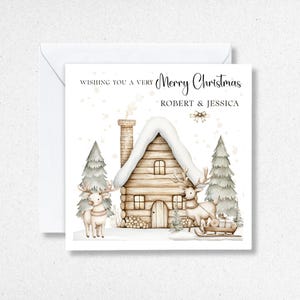 K&ouml;nnte beinhalten: Weihnachtskarte mit einer Aquarellillustration eines schneebedeckten Blockhauses, zwei Rentieren und Tannenb&auml;umen. Die Karte tr&auml;gt die Aufschrift "Wishing you a very Merry Christmas" und die Namen "ROBERT & JESSICA".