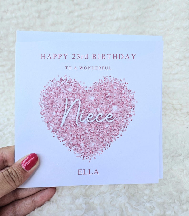 Może przedstawiać: Biała kartka z r&oacute;żowym brokatem w kształcie serca. Na kartce nadrukowany jest tekst "Happy 23rd Birthday to a wonderful Niece Ella".