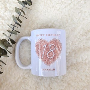 Może przedstawiać: Biały kubek ceramiczny z r&oacute;żowym sercem z numerem 18. Na kubku nadrukowany jest tekst "Happy Birthday Hannah".