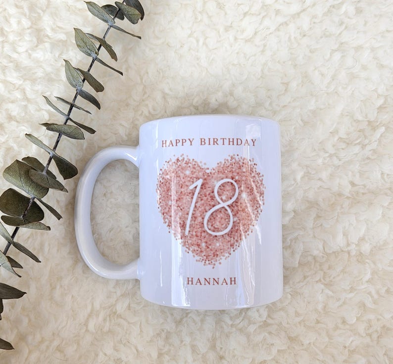 Może przedstawiać: Biały kubek ceramiczny z r&oacute;żowym sercem z numerem 18. Na kubku nadrukowany jest tekst "Happy Birthday Hannah".