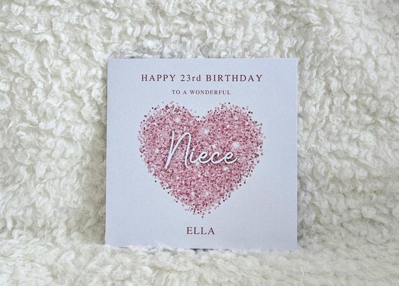 Może przedstawiać: Kartka z r&oacute;żowym sercem z brokatem, w kt&oacute;rym znajduje się napis "Niece" w białym kolorze. Na kartce widnieje tekst "Happy 23rd Birthday to a Wonderful Niece Ella".
