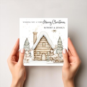 K&ouml;nnte beinhalten: Weihnachtskarte mit einer Aquarellillustration eines schneebedeckten Blockhauses, zwei Rentieren und Tannenb&auml;umen. Die Karte tr&auml;gt die Aufschrift "Wishing you a very Merry Christmas" und die Namen "ROBERT & JESSICA".