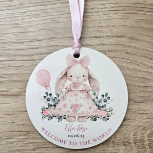Puede incluir: Un adorno redondo y blanco para anuncio de beb&eacute; con una cinta rosa. Presenta una ilustraci&oacute;n de un conejito con un vestido floral, un globo rosa y el texto "Welcome to the World". Tambi&eacute;n incluye el nombre y la fecha.