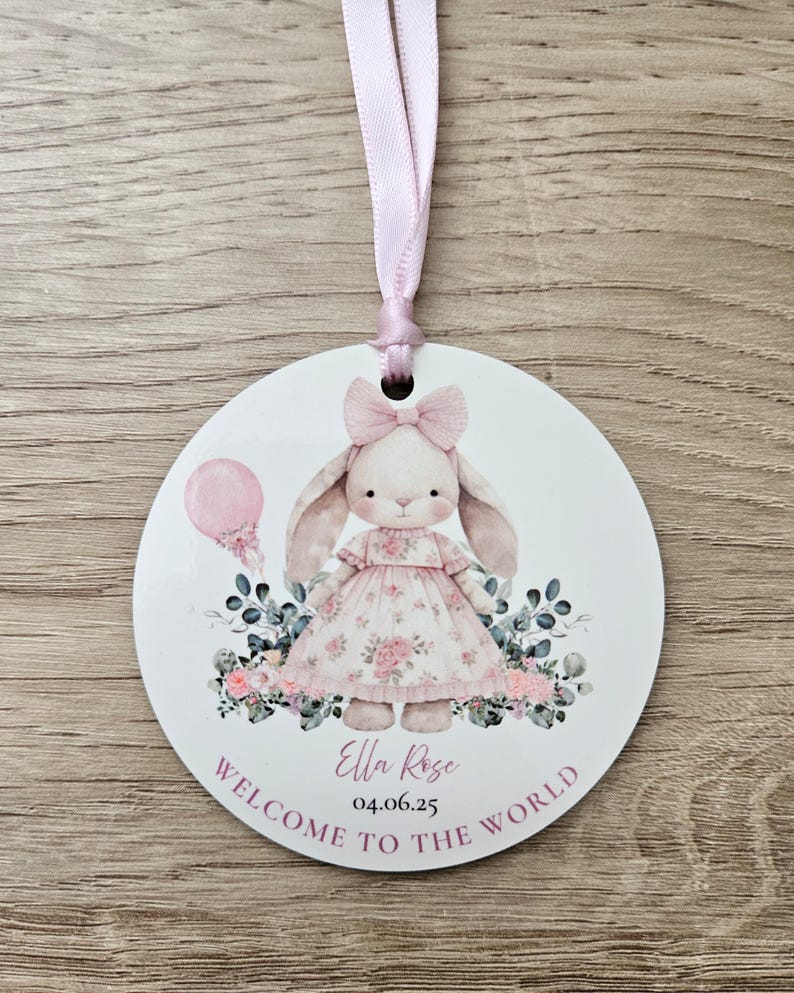 Puede incluir: Un adorno redondo y blanco para anuncio de beb&eacute; con una cinta rosa. Presenta una ilustraci&oacute;n de un conejito con un vestido floral, un globo rosa y el texto "Welcome to the World". Tambi&eacute;n incluye el nombre y la fecha.