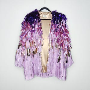 May include: A striking open-front jacket with a cascade of shimmering fringe in shades of purple, pink, and gold. The jacket's interior is a soft, champagne colour. The jacket is hanging on a black hanger.