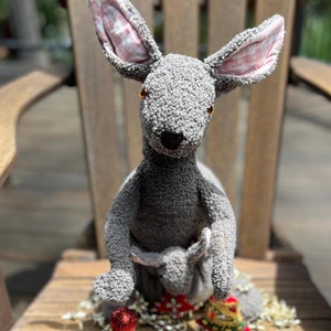 Puede incluir: Un juguete de peluche gris en forma de canguro con orejas rosas y un pequeño canguro en su bolsa. El canguro está sentado en una silla de madera con un adorno navideño rojo y dorado.