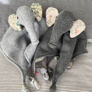 Könnte beinhalten: Mehrere graue Plüsch-Elefanten-Babyspielzeuge mit gemusterten Ohren und geknoteten Schwänzen. Die Ohren weisen Blumen- und Pflanzenmuster in verschiedenen Farben auf. Die Spielzeuge sind auf einem gestreiften Stoffhintergrund angeordnet.