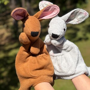 Puede incluir: Dos marionetas de mano de canguro. Una es marrón con el interior de las orejas rosa, la otra es gris con un estampado floral en el interior de las orejas. Ambas tienen ojos y nariz negros, y un bolsillo en la parte delantera.