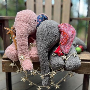 Peut inclure: Trois éléphants en peluche décorés pour les fêtes. Les éléphants sont rose, gris et bleu. Ils portent un tissu rouge et blanc à motifs autour du cou et sont ornés de guirlandes d'étoiles dorées.