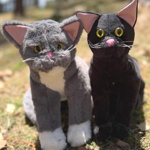 May include: Two plush cat toys. One is gray with white accents and pink ears, the other is black with pink accents. Both have yellow eyes and white whiskers. They are sitting on grass.
