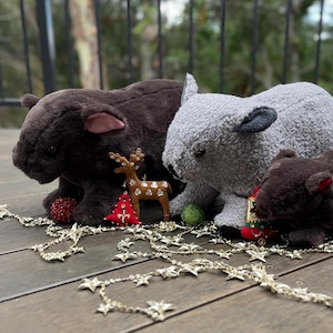 May include: Three plush toy wombats, one brown, one gray, and one dark brown, are sitting on a wooden floor with a gold garland of stars. The gray wombat is sitting in the middle and the other two are on either side of it. There are also small Christmas ornaments, including a red and white Christmas tree, a red ball, and a brown reindeer with rhinestones.